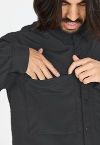 Zwarte button-up shirt gemaakt van lichtgewicht stof, met een ritszak op de borst en een grote voorkant zak voor opslag.