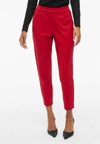 Pantalons tailleurs rouges avec une texture lisse, longueur cropped et plis à l'avant, associés à des escarpins noirs à bout pointu et un haut noir à motifs.