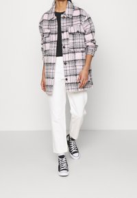 Chaqueta de camisa de cuadros en tonos de rosa, gris y negro, con un corte holgado, cierre de botones y dos bolsillos frontales. Combinada con pantalones blancos y zapatillas negras.