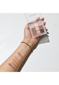 Oogschaduwpalet met negen vakjes in een doorzichtige behuizing. Swatches op de arm tonen tinten in beige, bruin en glitterafwerkingen. Gouden armband zichtbaar.