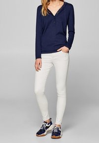 Navyblaues Langarm-Hoodie mit V-Ausschnitt und Knöpfen, kombiniert mit weißen, taillierten Hosen und navyblauen Sneakers mit braunen Akzenten.