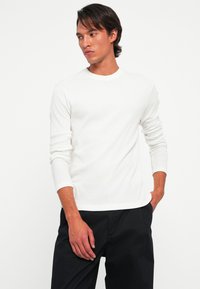 Man in een wit overhemd met lange mouwen en zwarte broek, staand met één hand in de zak, kijkend naar de zijkant tegen een witte achtergrond.