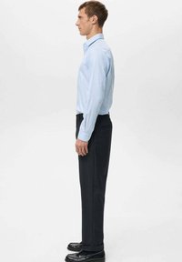 Svjetloplava prugasta košulja s ovratnikom na kopčanje, u kombinaciji s tamnim hlačama i crnim cipelama, prikazana iz profila.