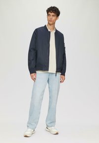 Marine bomberjack met een geribbelde kraag, ritssluiting aan de voorkant en zakken aan de zijkant, gedragen over een lichtkleurig T-shirt, gecombineerd met lichtblauwe jeans en witte sneakers.