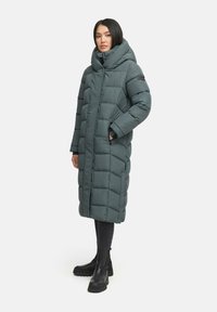 Long, puffy coat in dark teal, featuring a large hood, zip front, and quilted pattern. Worn with black ankle boots and slim dark pants.