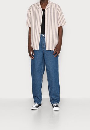 Camisa de manga corta a rayas en beige claro y lavanda, combinada con unos vaqueros de denim azul de corte holgado y unas zapatillas en blanco y negro.