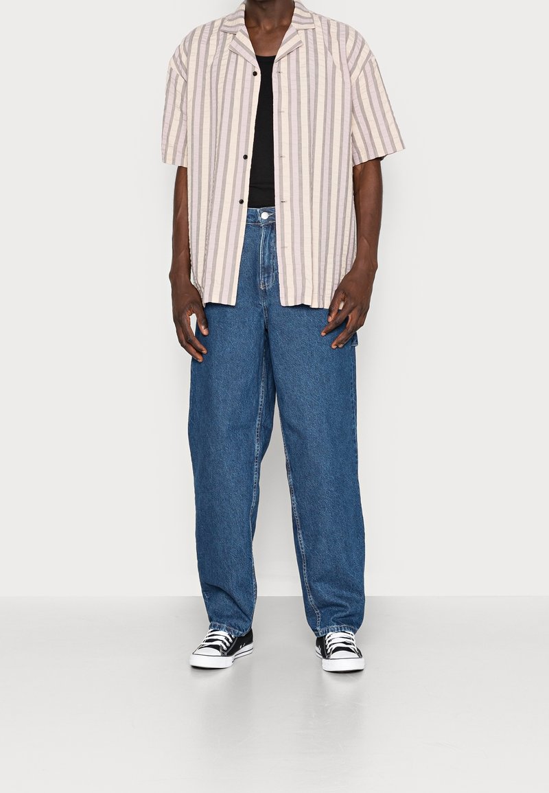 Lyhythihainen raidallinen paita vaaleanbeigeä ja lavenidia, yhdistettynä löysiin sinisiin farkkuihin ja musta-valkoisiin tennareihin.