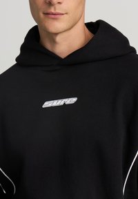 Giovane che indossa una felpa nera con bordi bianchi e un logo bianco centrato che recita "sure" sul petto.