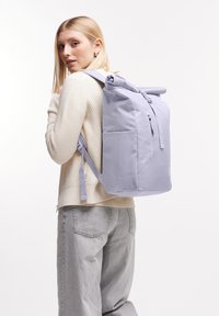 Vaaleanlila rullaläppäreppu, jossa on etupuolen vetoketjullinen tasku, säädettävät hihnat ja sileä kangas. Kuljetaan selässä.