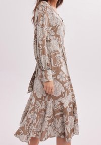 Femme portant une robe midi beige à manches longues avec des motifs floraux blancs, debout devant un fond clair uni.