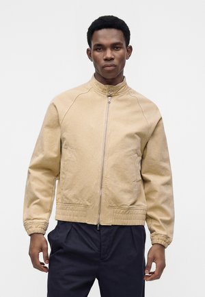 Mand med kort krøllet hår iført beige lynlåsjakke med høj krave og elastiske manchetter, parret med mørke bukser, stående mod hvid baggrund.