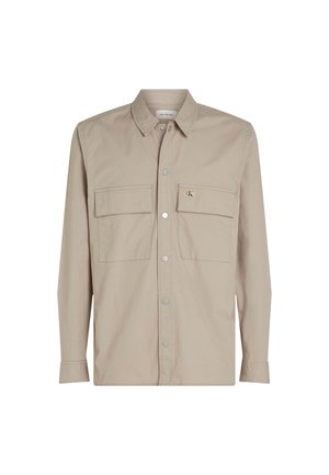 Camisa beige de botones con mangas largas, dos bolsillos en el pecho y un parche de logo en el bolsillo izquierdo. Tejido suave con cuello.