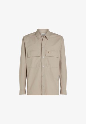 Camisa beige de botones con mangas largas, dos bolsillos en el pecho y un parche de logo en el bolsillo izquierdo. Tejido suave con cuello.