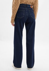 Jeans larghi di colore blu scuro con vita alta, dotati di una patch in pelle beige sul retro e tasche posteriori. Abbinati a un maglione a trecce beige.