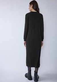 Dunkelgrünes, langärmliges Strickkleid mit geradem Schnitt, Rundhalsausschnitt und seitlichen Schlitzen, kombiniert mit schwarzen Stiefeletten.