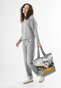 Stofftasche mit schwarz-weißer kariertem Muster, mit einem Design eines gelben Autos und Bergen. Grauer Pullover und Jeans, die von einem Modell getragen werden.