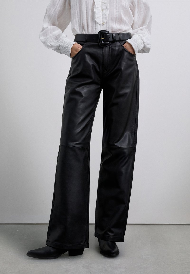 Zwarte leren broek met hoge taille, wijde pijpen en zijzakken, gecombineerd met een zwarte riem en aansluitende enkellaarzen.
