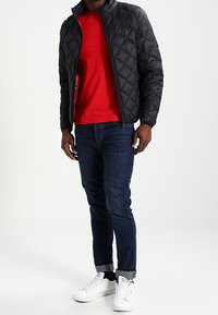 Schwarze gesteppte Jacke mit hohem Kragen, rohem Hemd darunter, dunkle Jeans und weiße Sneaker. Verfügt über eine Reißverschlusstasche an der Jacke.