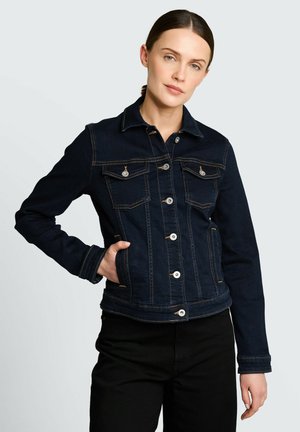 Frau mit dunklem Haar, die eine dunkelblaue Jeansjacke mit durchgehendem Knopfverschluss und Brusttaschen trägt, steht mit einer Hand in der Tasche vor einem einfarbigen Hintergrund.