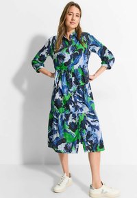 Blau- und grün gemustertes, knopfbares Kleid mit einem schichten Design und dreiviertel Ärmeln. Kombiniert mit hellen Sneakers.
