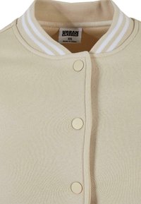 Beige jerseyjakke med hvitstripet ribbekrage, trykknapplukking og strukturert stoff. Merket sier "URBAN CLASSICS," størrelse XS.