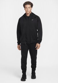 Zwarte sporttracksuit met een zip-up jas met textuurpanelen en een fitted ontwerp, gecombineerd met bijpassende broeken en schoenen.