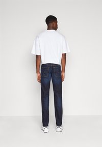 Hombre vestido con una camiseta blanca, jeans azul oscuro y zapatillas blancas, de pie frente a una pared gris claro lisa con las manos relajadas a los lados.