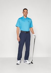Îmbrăcăminte de golf care include un polo bleu deschis cu puncte închise, pantaloni bleumarin, o curea neagră și pantofi de golf albi cu accente bleue, ținând un club de golf.
