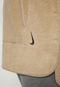 Beżowa bluzą polarowa o miękkiej, teksturowanej powierzchni, z czarnym logo Nike w kształcie swisha oraz beżowym wykończeniem wzdłuż krawędzi i kieszeni.
