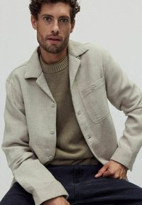 Ljust beige linneknapp-jacka med en bröstficka, lagd över en strukturerad olivgrön tröja, kombinerad med mörka jeans.