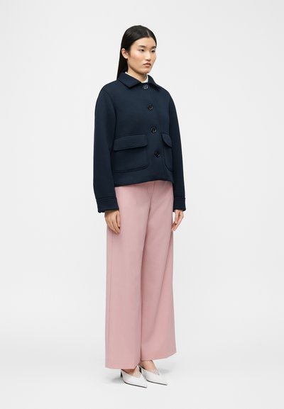 Γυναίκα που στήνεται φορώντας ένα ναυτικό μπλε σακάκι με κουμπιά και τσέπες με καπάκια, ροζ παντελόνι φαρδύ στο πόδι και άσπρα τακούνια με μυτερή μύτη σε απλό φόντο.