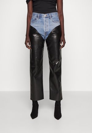 Osoba nosząca spodnie z wysokim stanem z niebieskiego denimu na górze i czarnej skóry na dole, w połączeniu z czarnymi butami w szpic.