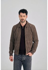 Mężczyzna o ciemnych włosach, ubrany w brązową zamszową kurtkę zapinaną na zamek, na czarnej koszulce i niebieskich dżinsach, stoi z jedną ręką w kieszeni na szarym tle.