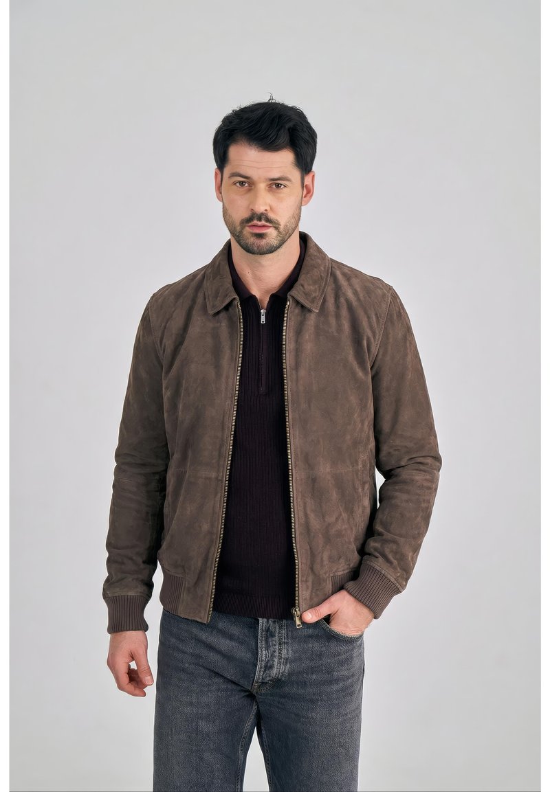 Mężczyzna o ciemnych włosach, ubrany w brązową zamszową kurtkę zapinaną na zamek, na czarnej koszulce i niebieskich dżinsach, stoi z jedną ręką w kieszeni na szarym tle.