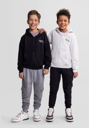 Dwóch uśmiechniętych chłopców, jeden w czarnej bluzie z kapturem i szarych spodniach bojówkach, drugi w białej bluzie z kapturem i czarnych spodniach bojówkach, stojących obok siebie.