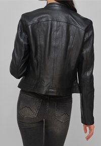 Donna che indossa una giacca di pelle nera aderente e jeans neri sbiaditi, vista da dietro su uno sfondo grigio semplice.