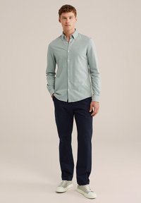 Bărbat în picioare, îmbrăcat cu o cămașă verde deschis cu nasturi, pantaloni negri închis și adidași albi, pe un fundal beige simplu.