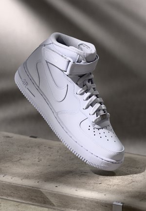 Witte hoge Nike sneaker met veters en enkelbandje, zwevend boven een gestructureerd stenen oppervlak met een zachte schaduw op de achtergrond.