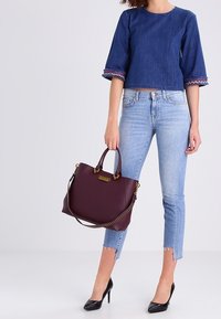 Borsa a tracolla bordeaux con doppi manici, dettagli dorati e finitura texture. Il modello indossa un top di jeans blu cropped e jeans strappati azzurri.