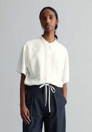 Woman with dark hair in a ponytail wearing a loose white button-up shirt and high-waisted black pants with a white drawstring on a gray background.