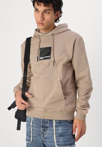 Beige hoodie med en sort firkantet logo lomme. Har snørebånd, en kengurulomme og ribbede manchetter. Båret med slidte blå jeans.