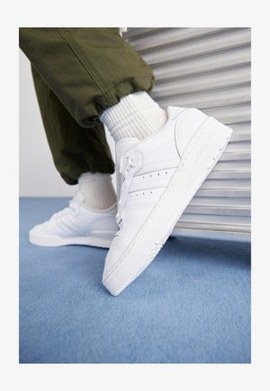 Valged tennisjalatsid, millel on sile nahkviimistlus, perforatsiooniga triibud, lapikud paelad ja tekstureeritud kummitald, koos oliivroheliste pükstega.