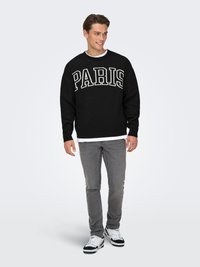 Black sweater with "PARIS" in white letters, gray jeans, and white sneakers. Crew neck design with ribbed cuffs and hem. Casual outfit.