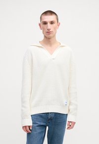 Kremfarget strikket genser med v-hals og ribbestrikkede mansjetter. Har et teksturert mønster og et logo-patch nær hemline. Båret med blå jeans.