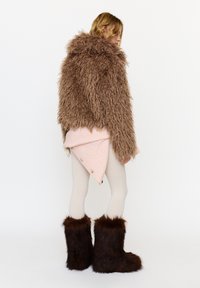 Kvinde iført en blød brun pelsjakke, lyserosa nederdel med knapper, cremefarvede leggings og mørkebrune pelsstøvler, stående mod en hvid baggrund.