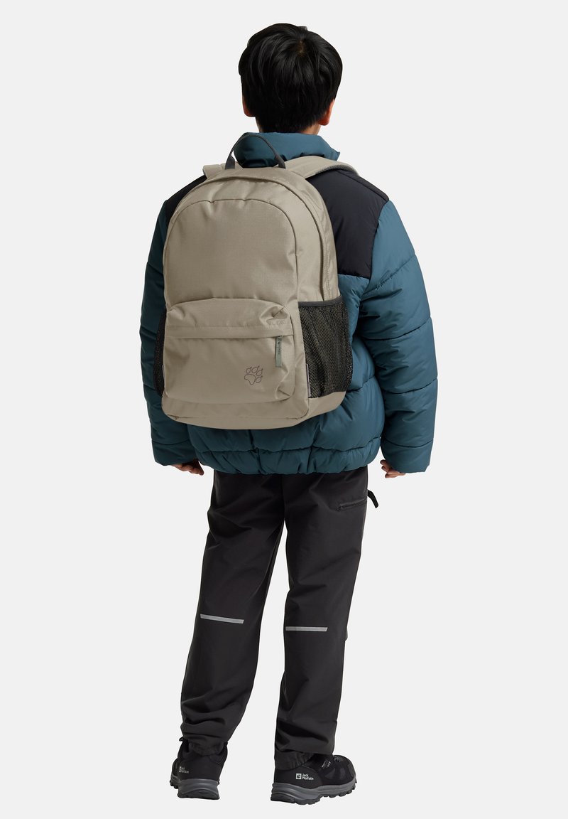 Sac à dos beige avec un compartiment principal, une poche avant et une poche latérale en mesh, porté sur une veste en duvet bleue et un pantalon noir.