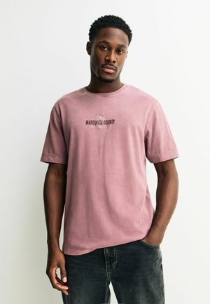 Fiatal férfi kopott rózsaszín Marquette megyei pólóban és sötét farmerral, egyik kezét zsebében tartva áll sima fehér háttér előtt.