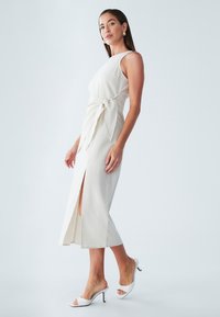 Vrouw draagt een mouwloze beige midi-jurk met zijsplit en ceintuur, gecombineerd met witte hoge hakken en gouden oorbellen.