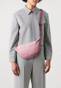 Kompaktní růžová manšestrová kabelka přes rameno s zakřiveným tvarem, nastavitelným popruhem a texturovaným povrchem, nošená přes černo-bílé kostkované oblečení.