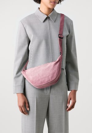 Kompakt pink skuldertaske i fløjl med en buet form, justerbar rem og tekstureret overflade, båret over et sort- og hvidternet outfit.